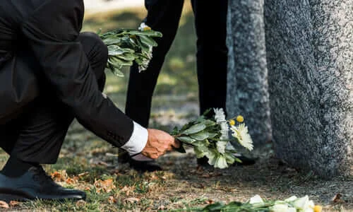 Two people in black mourning and leaving flowers at a deceased loved one's grave.