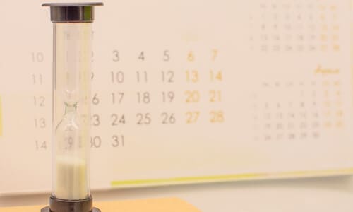 A closeup of a trickling hourglass in front of a calendar on a desk.