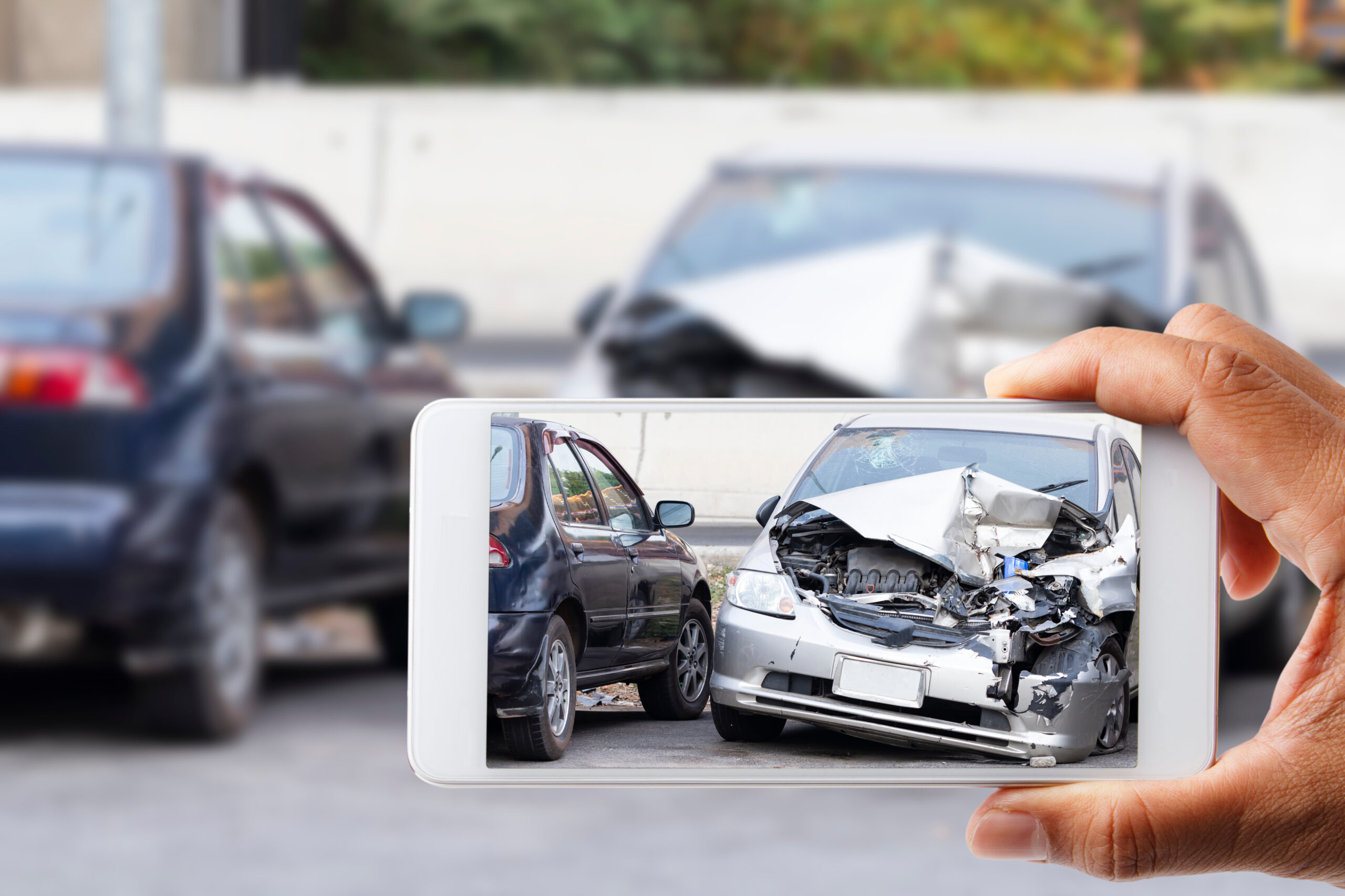 In the foreground, a hand holds a smartphone to capture a photo of the damage to their car after an accident.