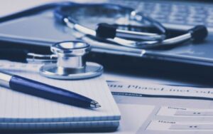 A close up of a stack of medical papers, a pen, a computer, and stethoscope.