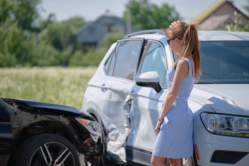 A frustrated woman leans against her white SUV after being hit by an uninsured driver in a black sedan.