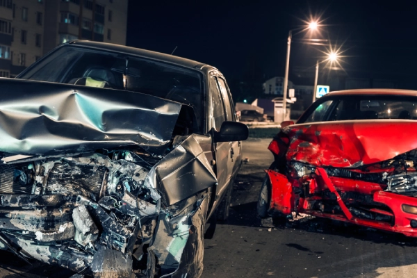 A nighttime photograph of a black car and a red car on an urban road after a head-on collision totaled them both.