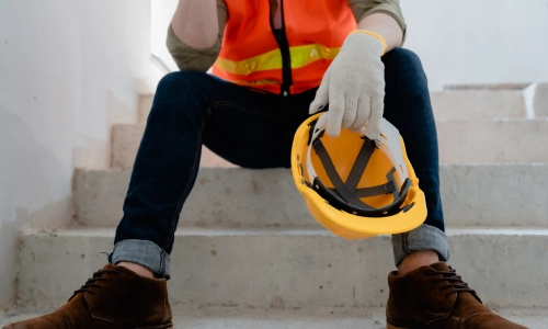 An injured construction worker sitting down on a staircase and holding his helmet while resting.