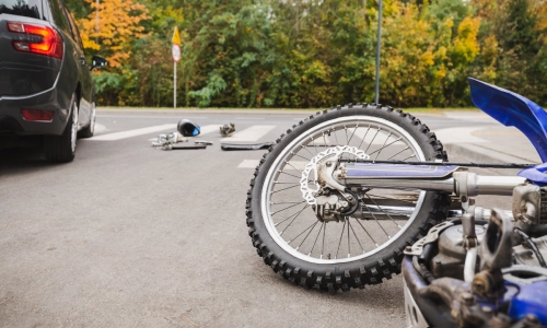 A collision between a motorcycle, a bicycle, and a black car on a suburban pedestrian crossing.