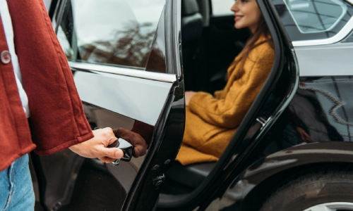 A rideshare driver opening the door for a passenger in a yellow coat to step out.