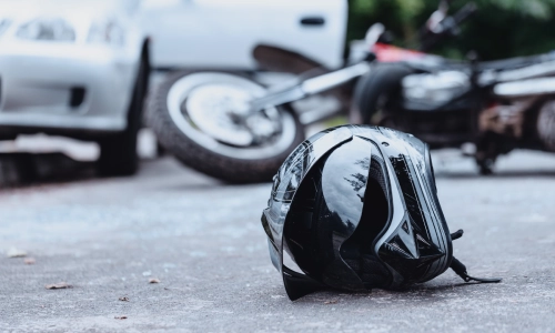 Close-up photo of a black biker helmet on the street with an overturned motorcycle in the background.