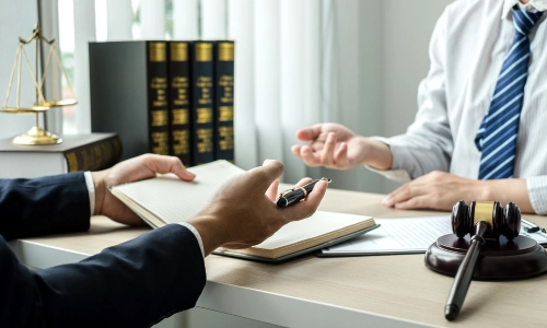 Two men are discussing legal matters with a gavel hammer on the table and a scale of justice in the background.