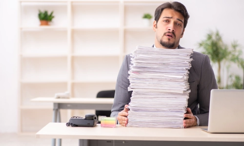 Unhappy young male employee sitting at a desk with piles of unresolved documents.